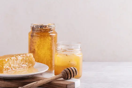 a bottle of chunk honey with a piece of honeycomb on a plate and a jar of raw behind a dipper